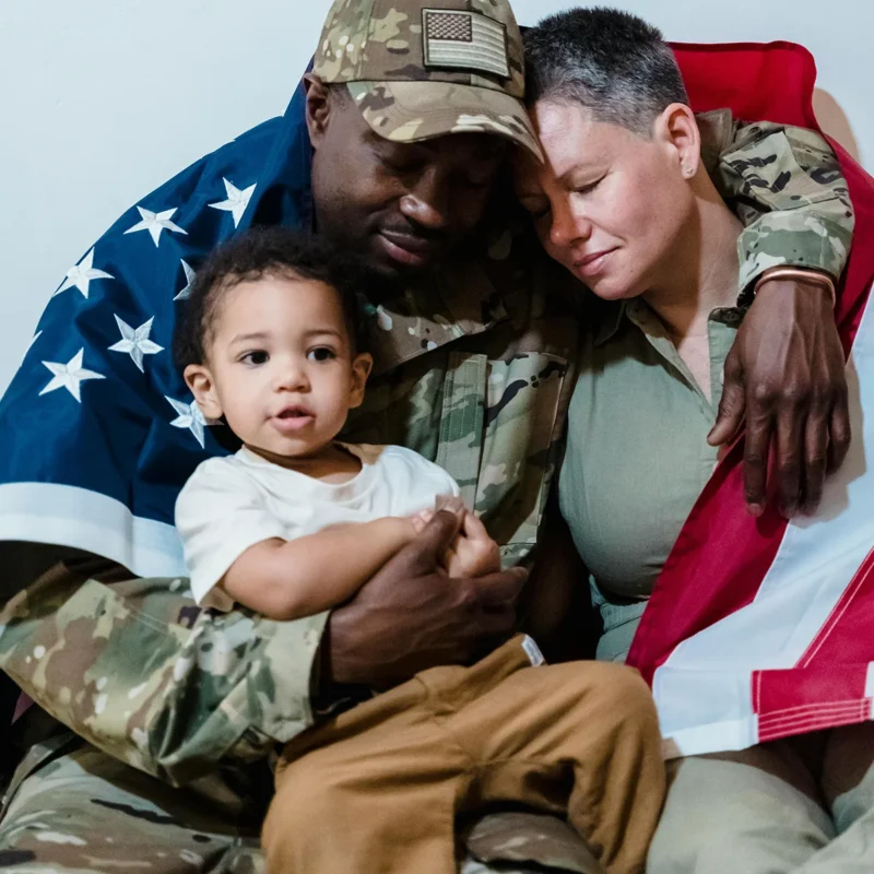 African American soldier hugging family with flag around back.